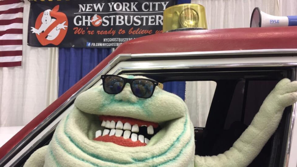 Slimer puppet wearing sunglasses, sitting inside of an Ecto 1 replica, in front of NYC Ghostbusters sign.